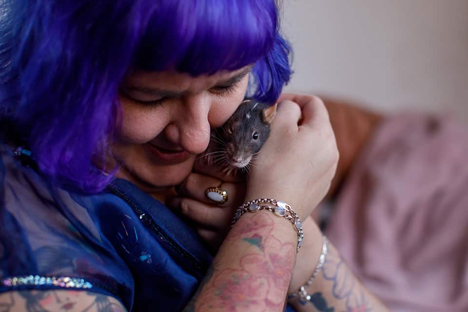 Vrouw met paars haar en tatoeages drukt ratje liefdevol tegen haar gezicht 