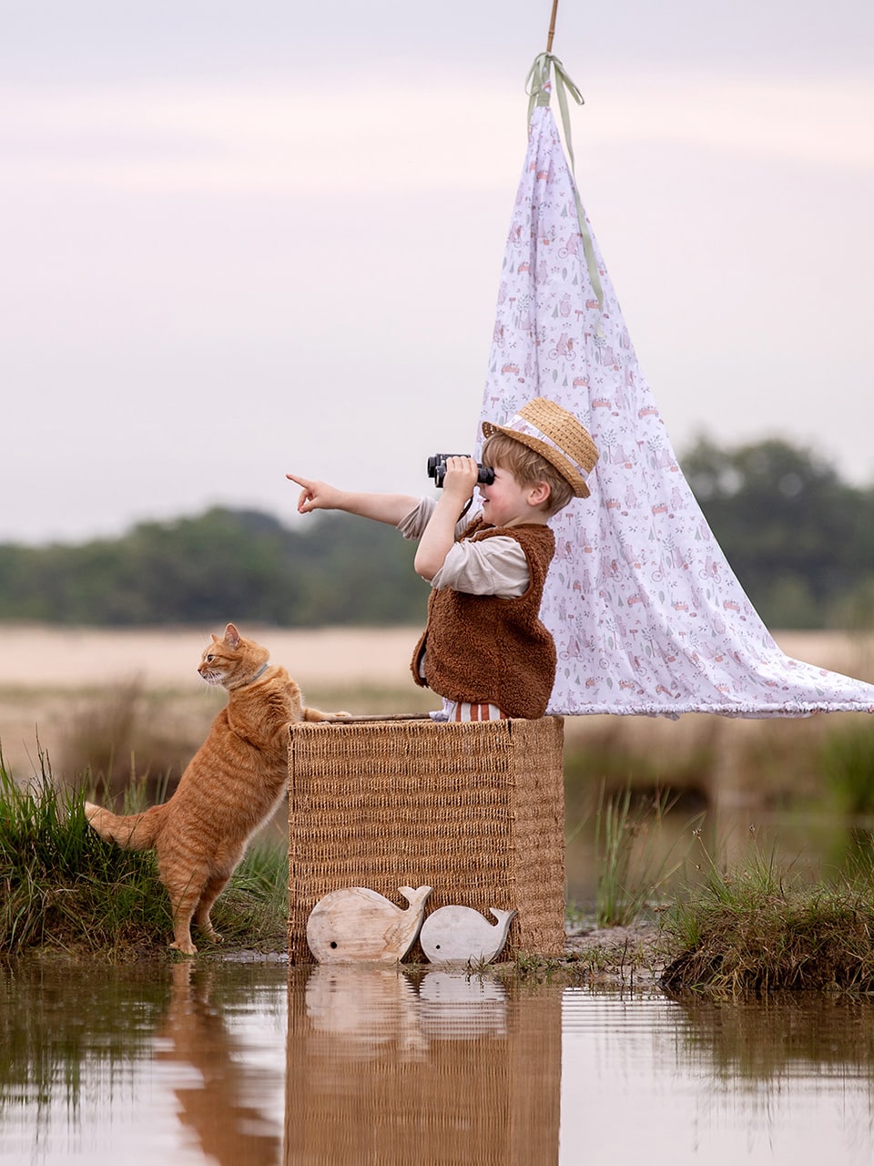 Jongen in zelf geknutselde zeilboot kijkt door verrekijker terwijl kat naast de zeilboot in de zelfde richting kijkt