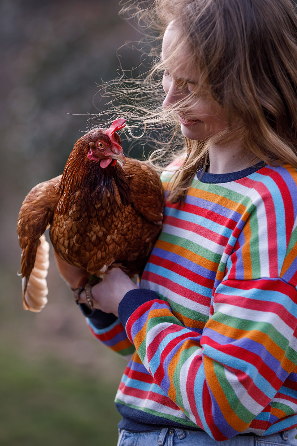 Jonge vrouw in bonte trui kijkt glimlachend naar kip op haar arm, terwijl het licht prachtig door haar haren schijnt. 