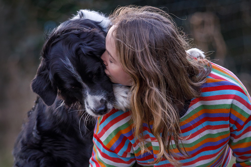 Jonge vrouw geeft hond die met zijn voorpoten over haar schouder hangt een kus. 