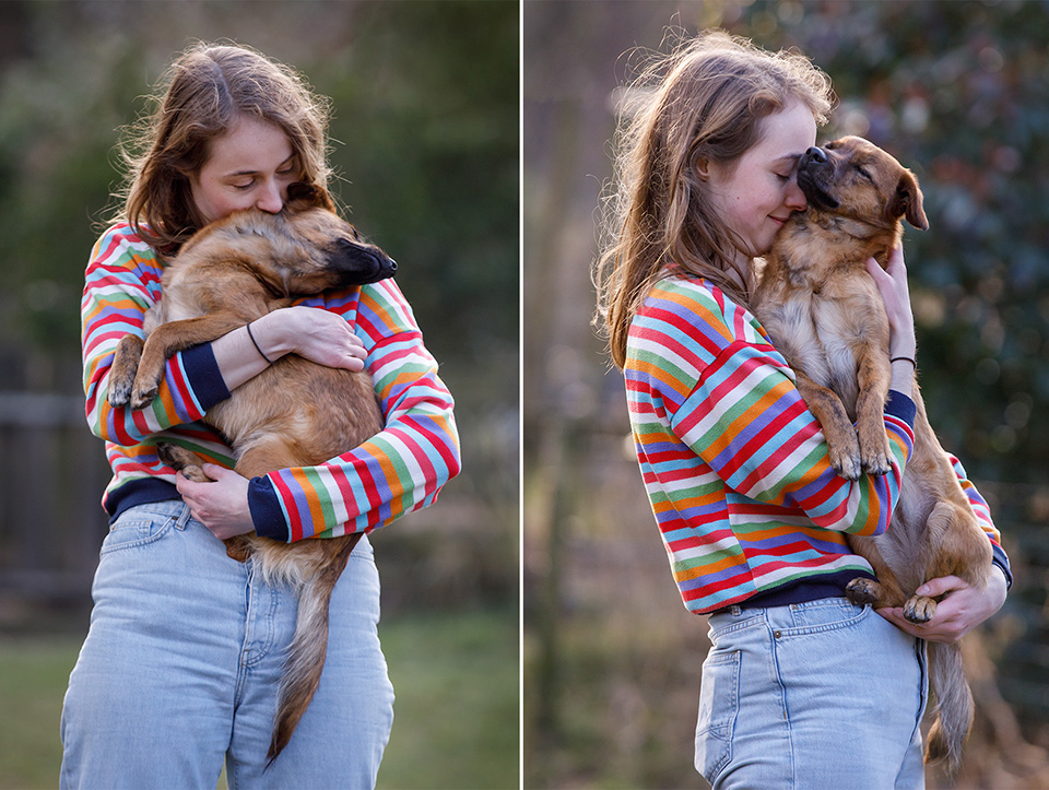 Jonge vrouw heeft bruin hondje op haar arm. Zij kust hem en hij vleit zich vol vertrouwen tegen haar aan. 
